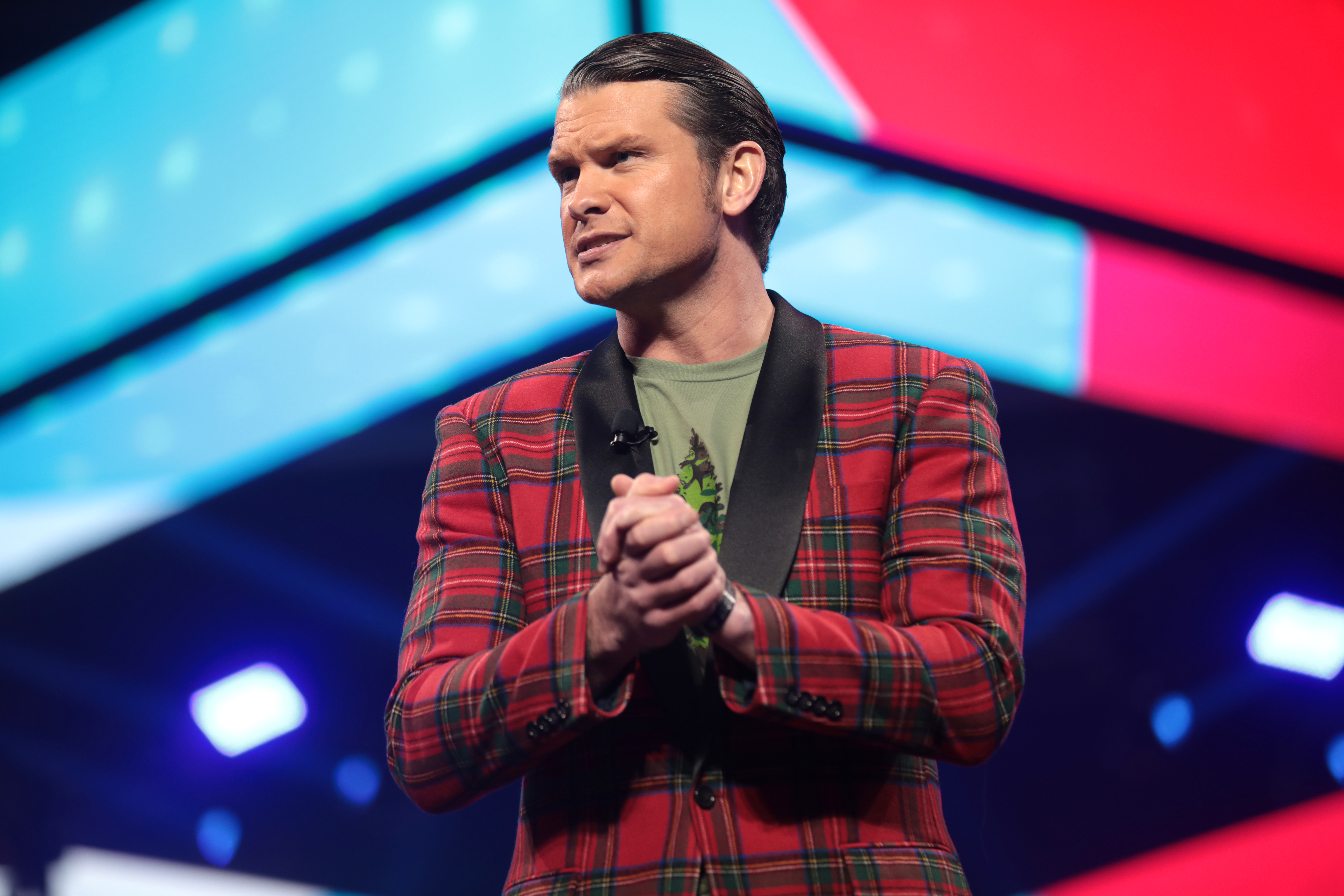 Pete Hegseth speaking at a public event in front of a bright stage backdrop.